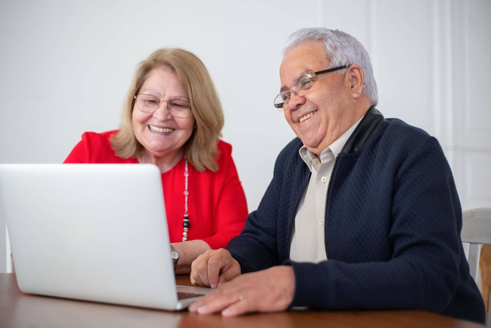 An older couple sitting at a table, smiling as they look at an open laptop.